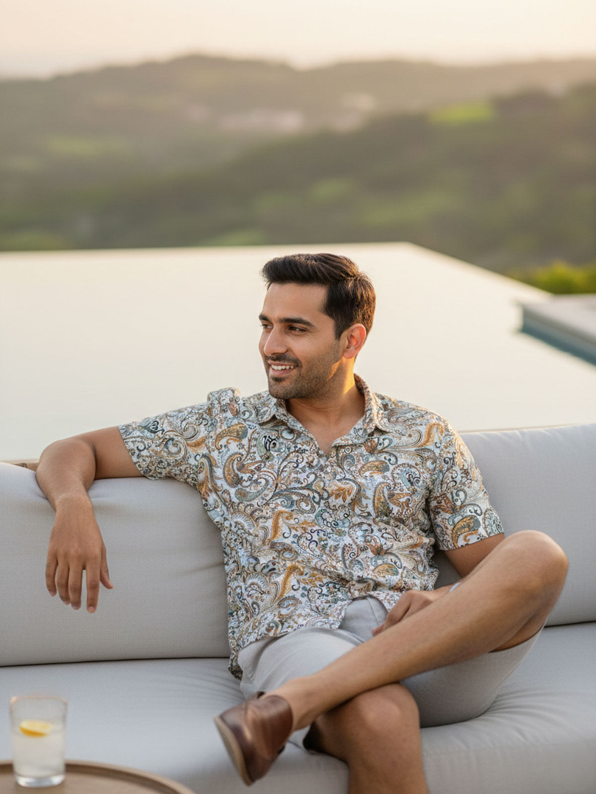 Man sitting on a white couch with a scenic background, wearing Privee Paris Paisley Print Shirt
